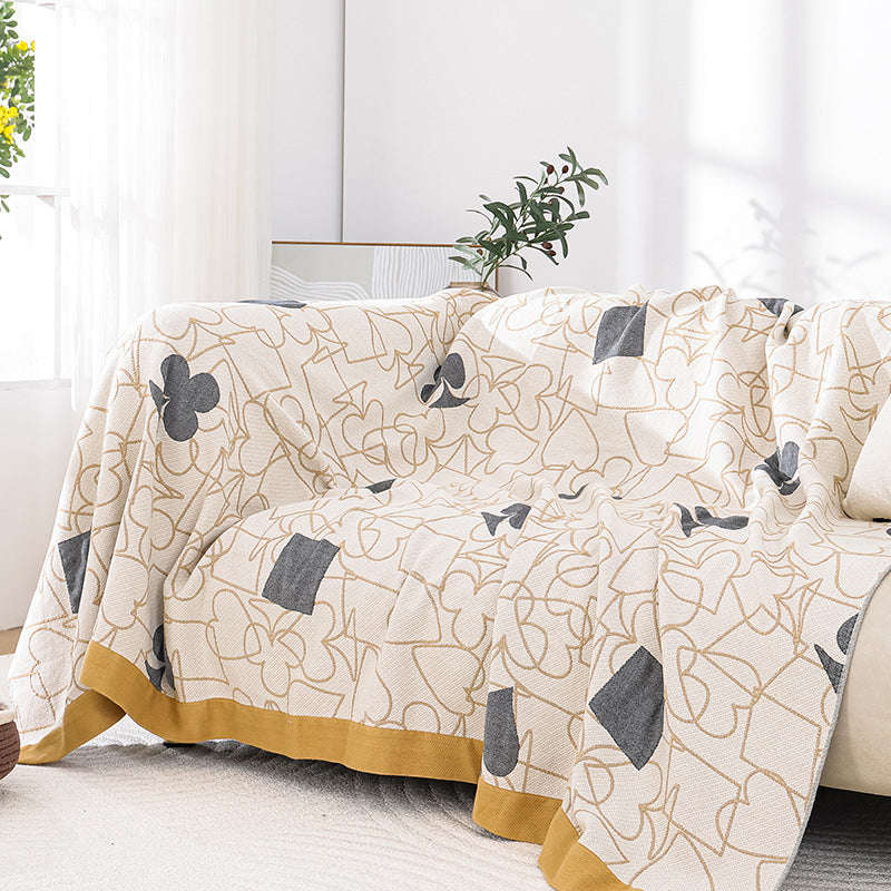 Cream card-suit jacquard gauze blanket draped over a light sofa, showing grey spades, hearts, clubs and diamonds with mustard border