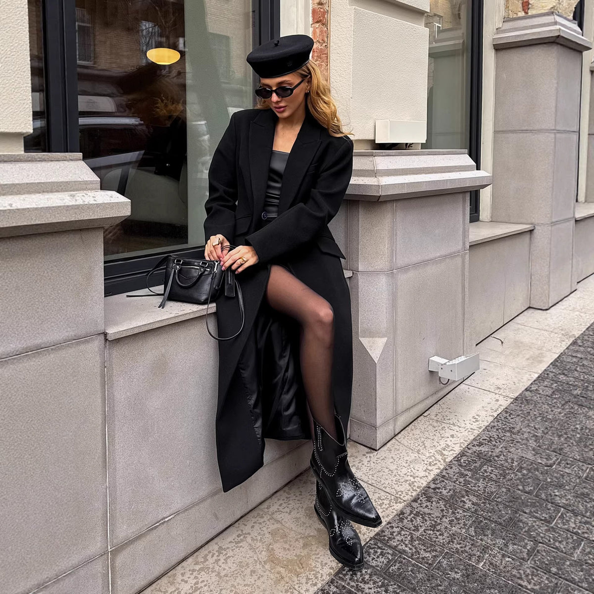Floor-length single-breasted tailored coat in black, worn by a model seated on a stone ledge, front view with beret and boots