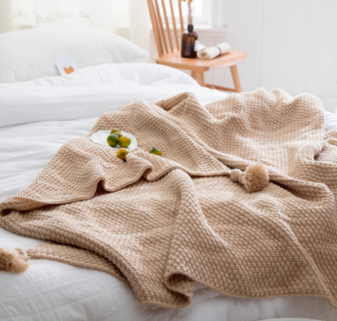 Beige chunky knit throw spread on a bed with pompoms and small fruit on a plate