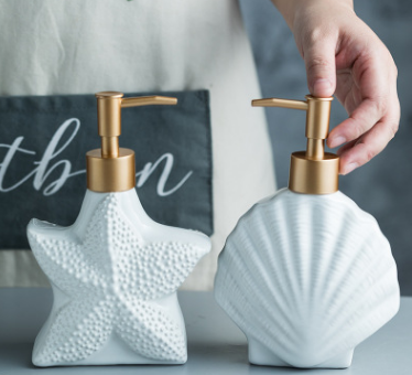 Ceramic starfish and shell soap dispensers with gold-tone pump tops, side by side on a surface, hand pressing the shell