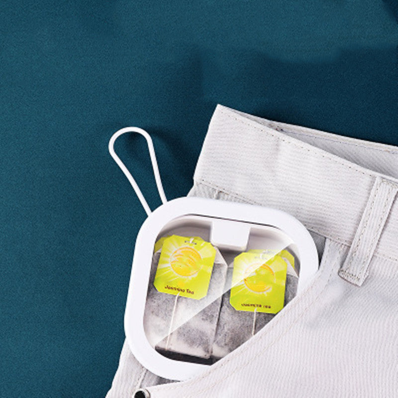 White storage case with tea bags inside, clipped onto the pocket of light grey trousers against a teal background