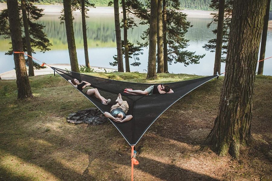 Three-point triangle hammock in black, suspended between trees near a lake, three people lying on it