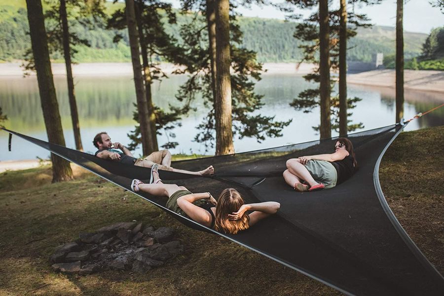 Three-point triangle hammock in black, hanging between trees by a lake, three people relaxing on it