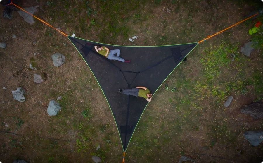 Three-point triangle hammock in black with green trim, viewed from above, two people lying on it on a grassy ground with
