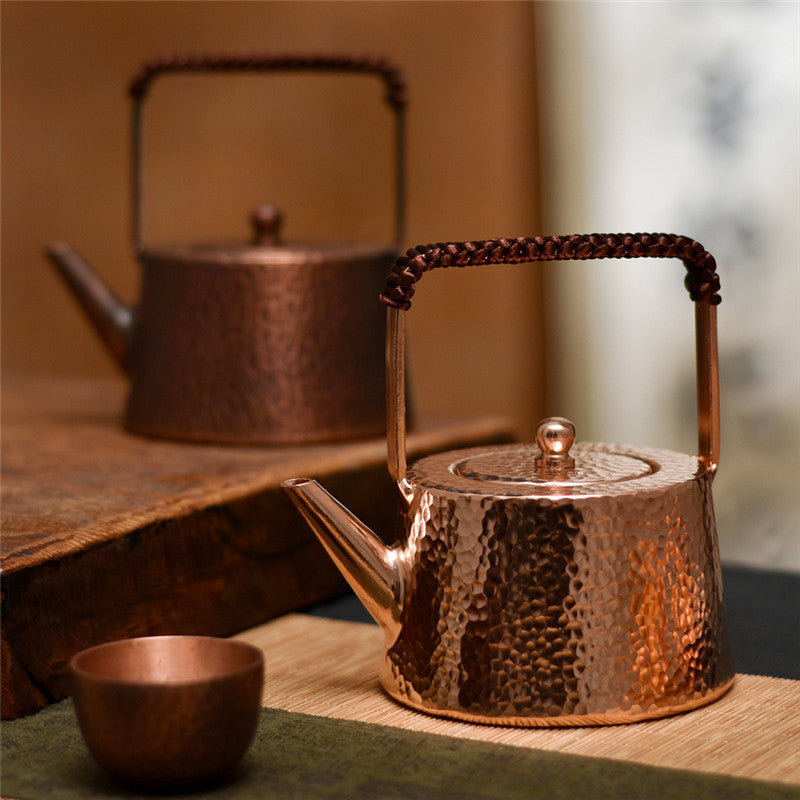 Hammered copper teapot with braided handle, front view on wooden surface, small cup nearby