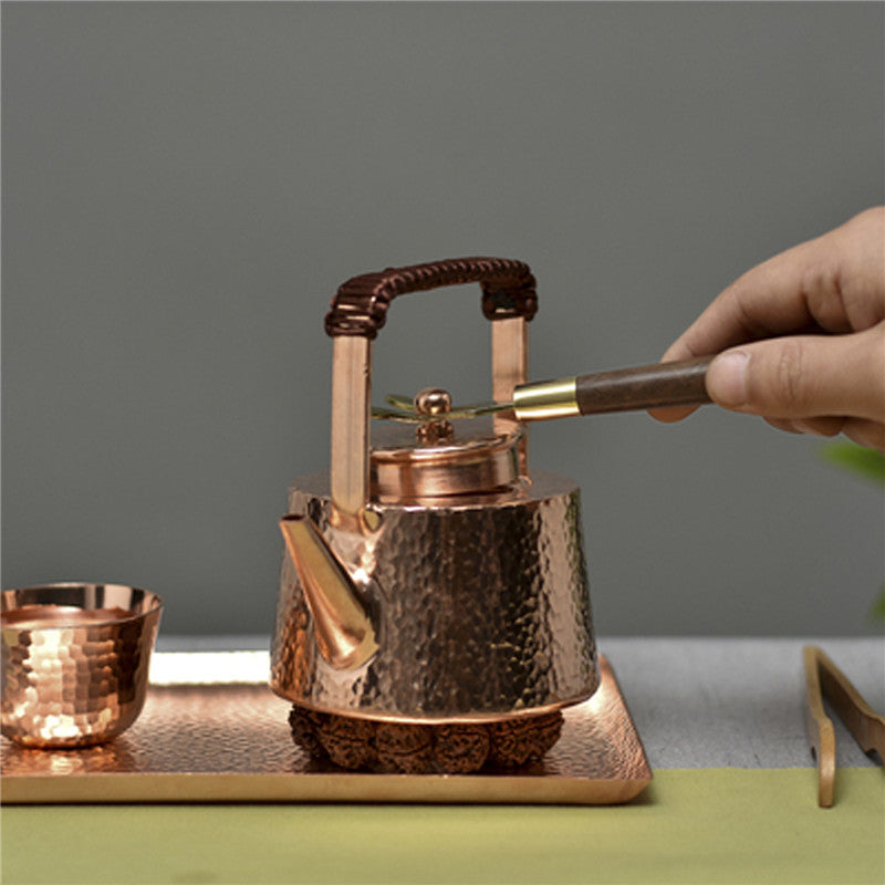 Hammered copper teapot with braided handle, in use, lid being opened with tool, tray and cup visible