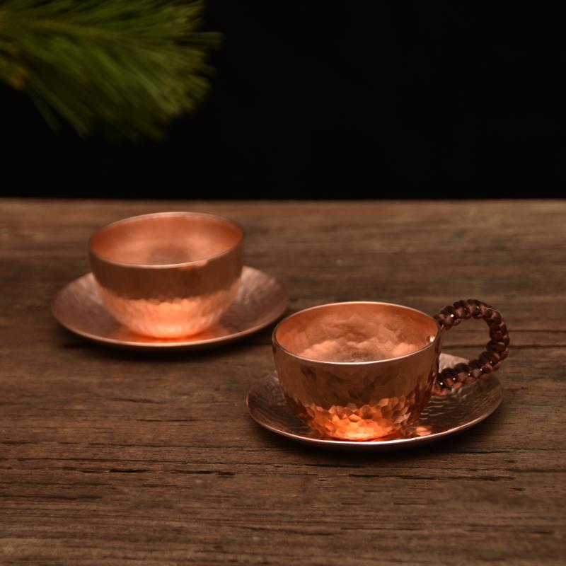 Two hammered copper-tone teacups with saucers, one with woven wire handle, on wooden surface