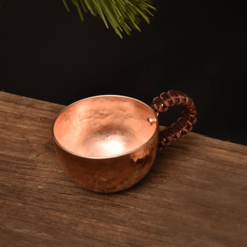Hammered copper-tone teacup with woven wire handle on wooden surface