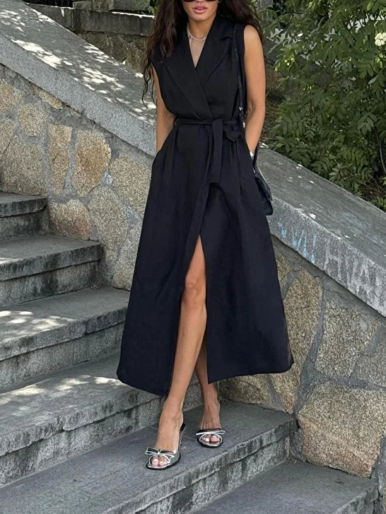 Sleeveless wrap blazer dress in black, front view on model, with tie belt and high slit, standing on stone steps