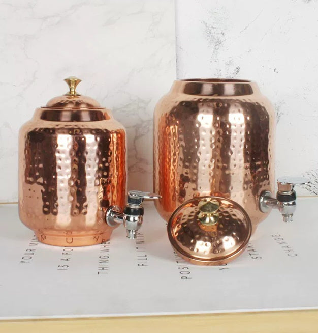 Two hammered copper-look water dispensers with tap, one lid removed, displayed on a flat surface with text background