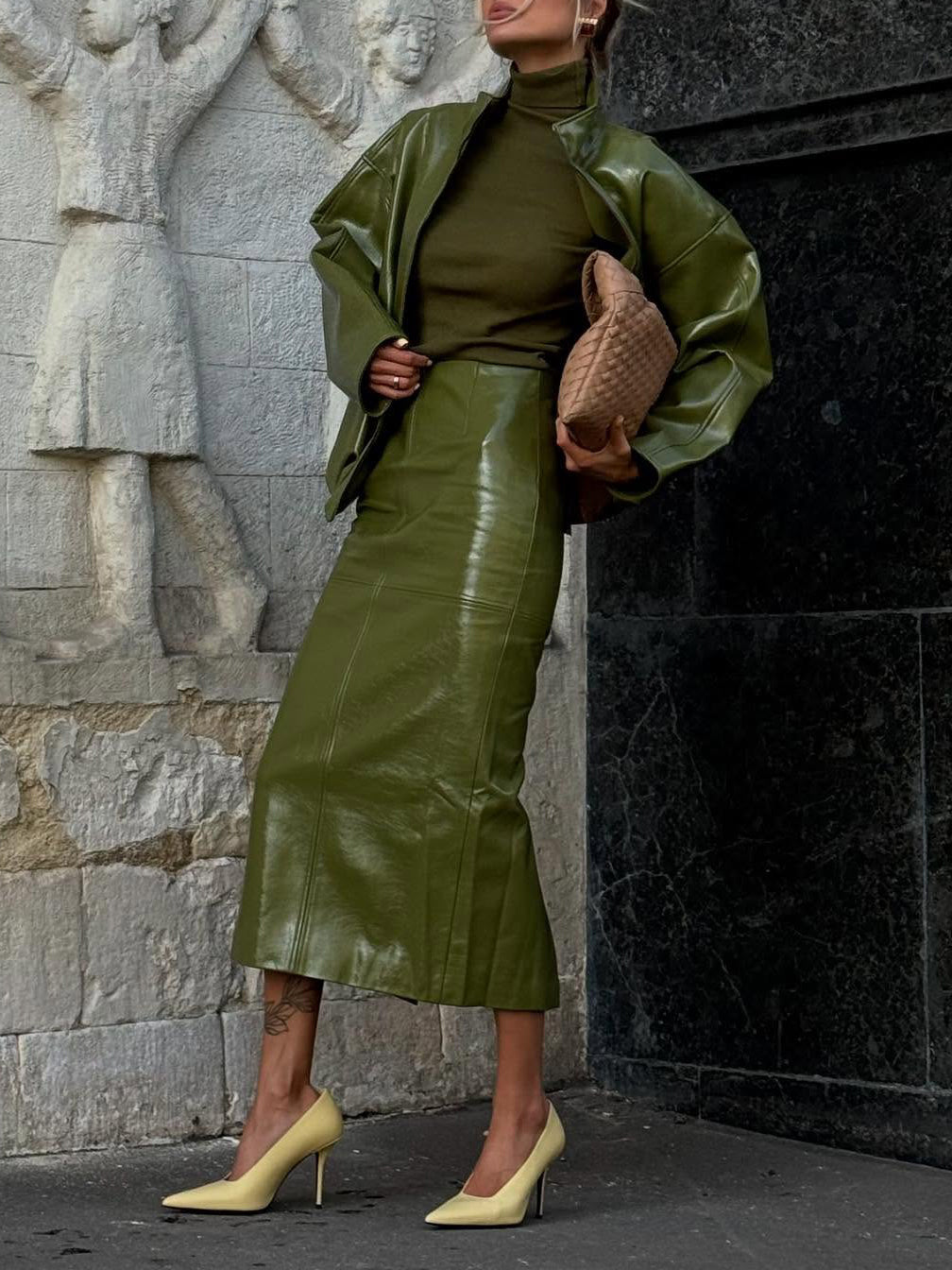 Faux-leather jacket and midi skirt co-ord set in olive green, front view on model, holding brown bag, standing near stone