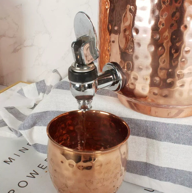 Close-up of hammered copper-look water dispenser tap pouring liquid into a matching cup on a striped cloth