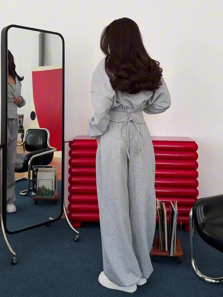 Grey corset top, cropped hoodie and wide-leg trousers set on model, back view, standing near mirror and red cabinet