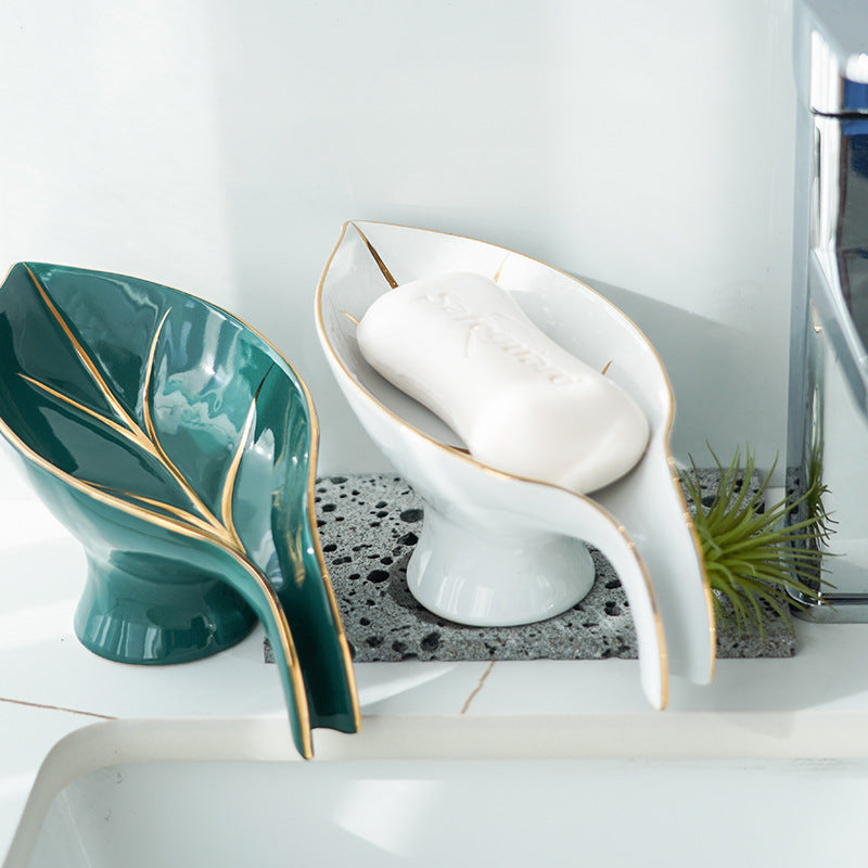 Green and white ceramic leaf-shaped soap dishes with gold-tone trim placed beside a sink