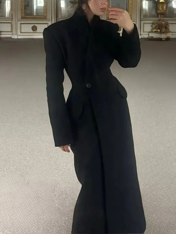 Model wearing a long black tailored coat with padded shoulders and a single waist button, standing indoors