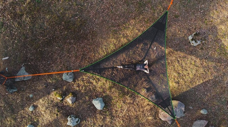 Three-point triangle hammock in black with green trim, aerial view, one person lying on it on dry ground