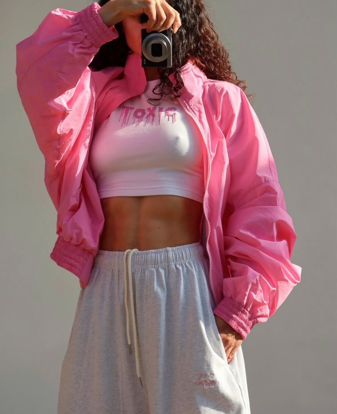 Bright pink cropped bomber jacket, front view on model, open, one arm raised, over white crop top and grey sweatpants