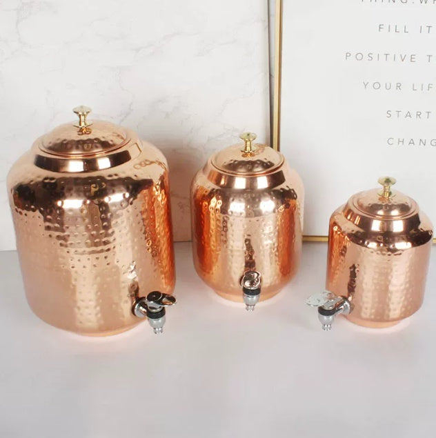 Three hammered copper-look water dispensers with tap, arranged in descending size on a white surface near a framed quote
