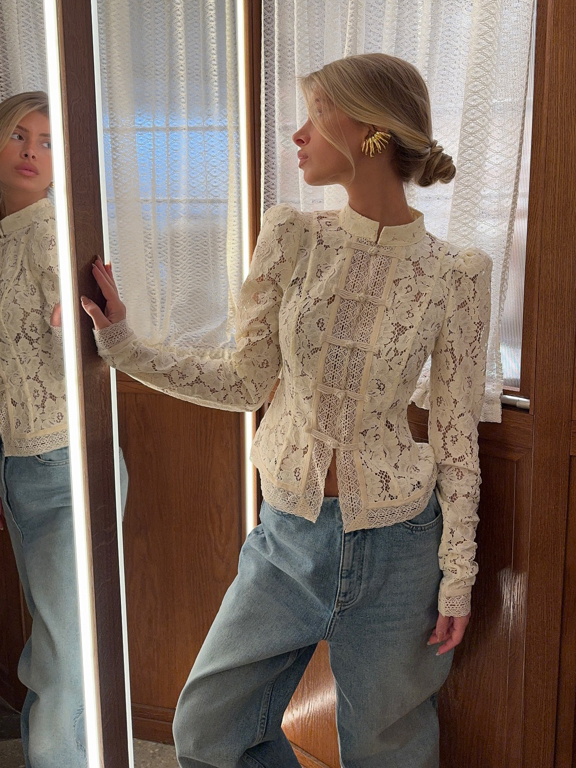 Side view of cream lace blouse with puffed shoulders and long sleeves, worn by model leaning against wooden frame