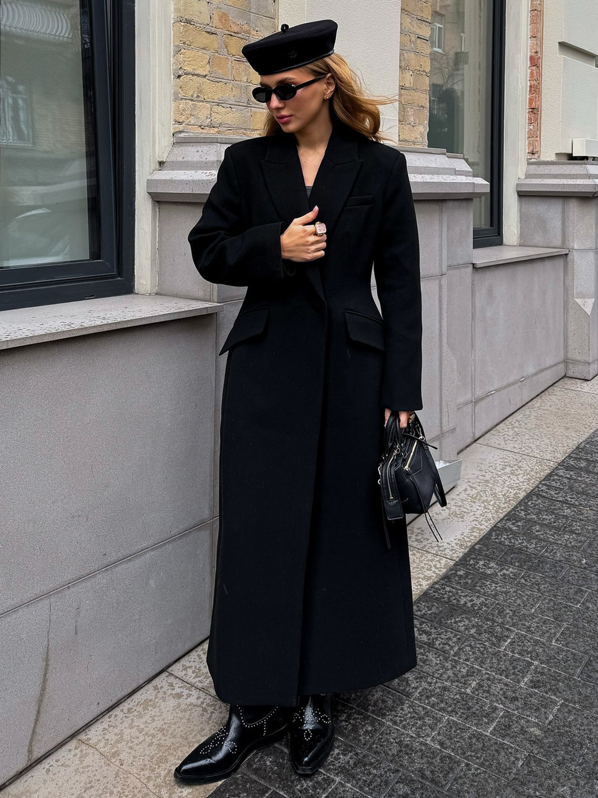 Floor-length single-breasted tailored coat in black, front view of model standing outdoors near a building