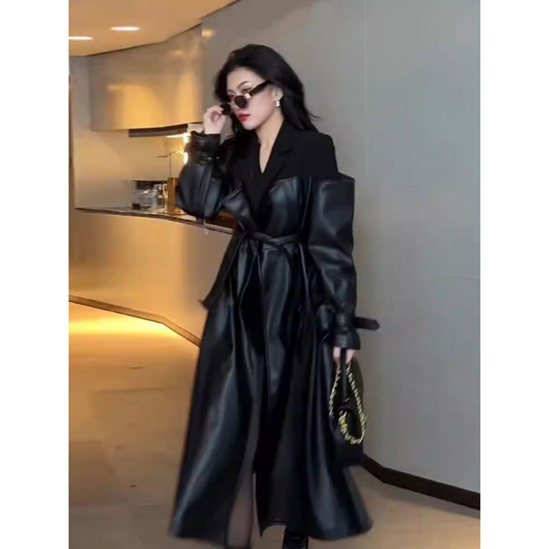 Model wearing long black trench coat with contrast shoulders, belt tied at waist, standing indoors