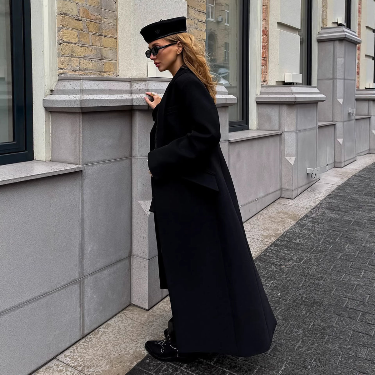 Floor-length single-breasted tailored coat in black, side view of model standing against a building wall