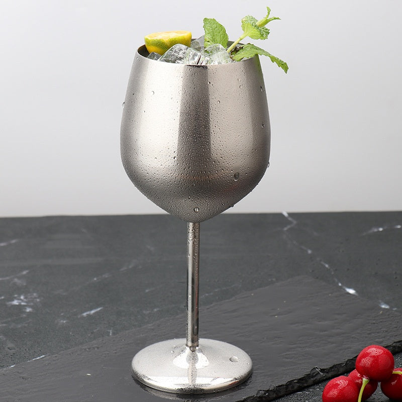 Stainless steel wine glass in silver, filled with ice, lemon slice, and mint, on dark surface with cherries nearby