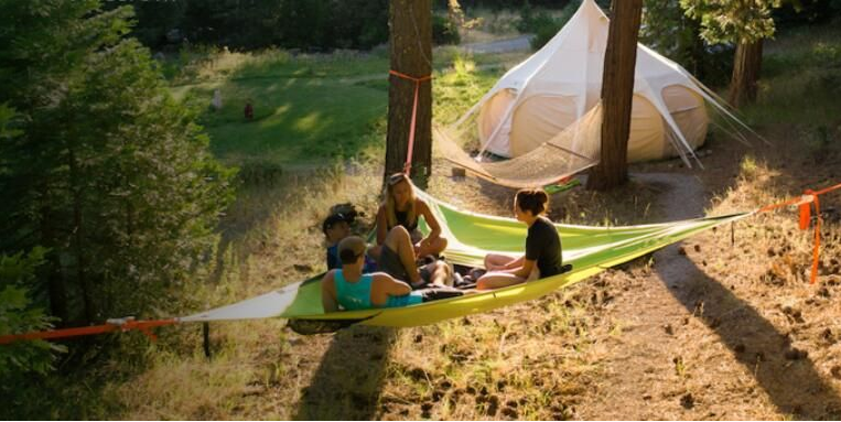 Three-point triangle hammock in green and white, set up between trees near a tent, four people sitting on it