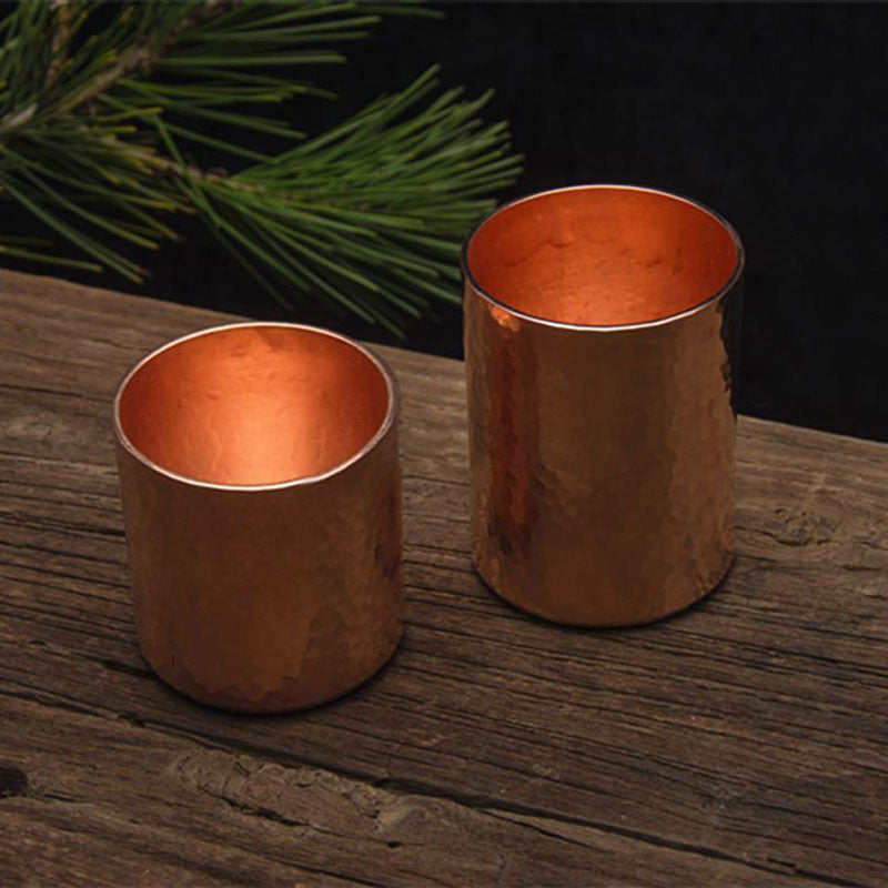 Two hammered metal tea cups, copper colour, placed on wooden surface with pine branch in background