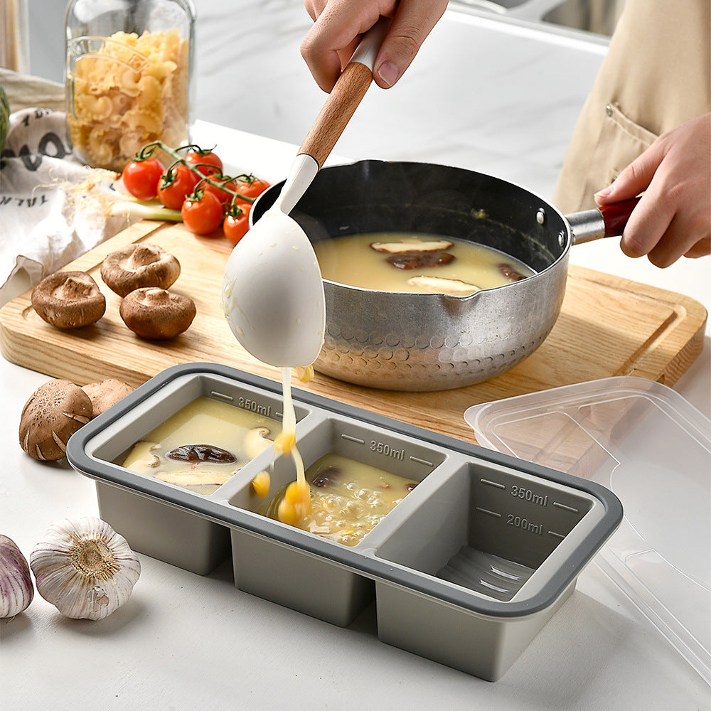 Silicone freezer portion tray with lid, three compartments, grey rim, measuring marks, being filled with soup in kitchen