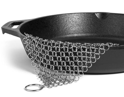 Stainless steel chainmail scrubber on a black cast iron pan, showing use in cleaning, side view, white background