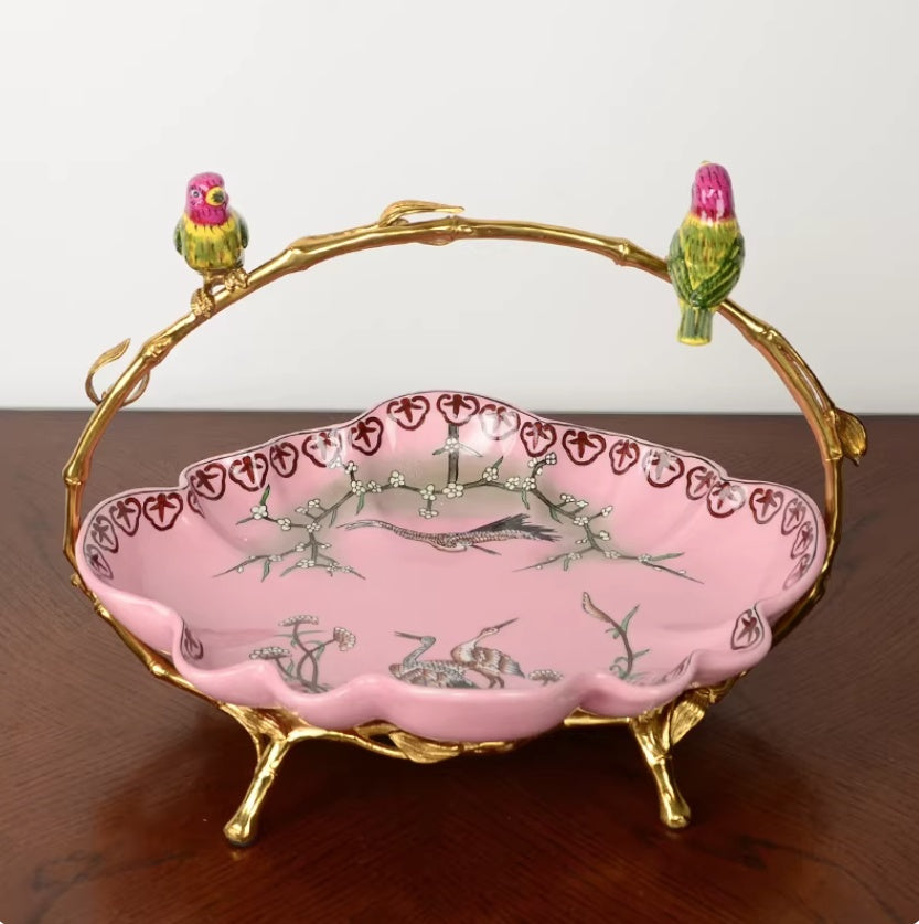 Pink ceramic fruit bowl with crane and branch motif, gilt branch handle and two birds, front view on wooden surface