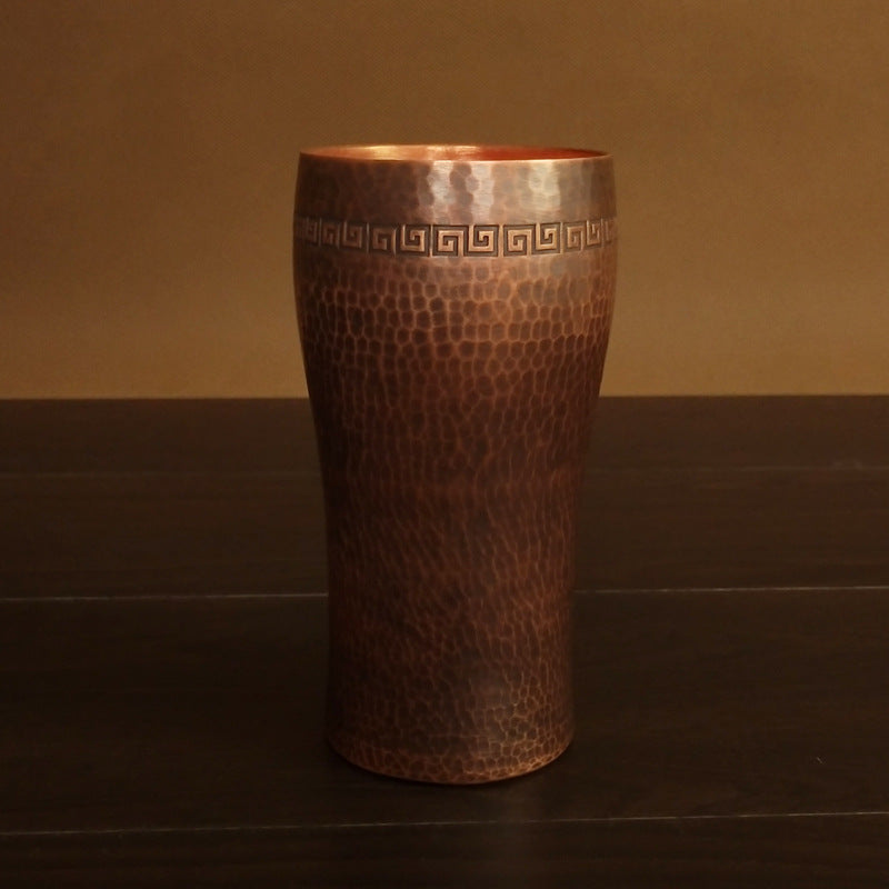 Curved hammered copper beer tumbler with Greek-key border, on dark surface