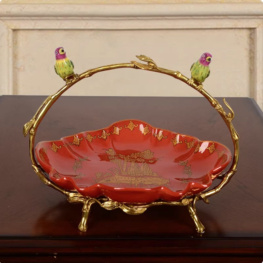 Red ceramic fruit bowl with gold pattern, scalloped edge, gilt branch handle with two birds, on dark wooden surface