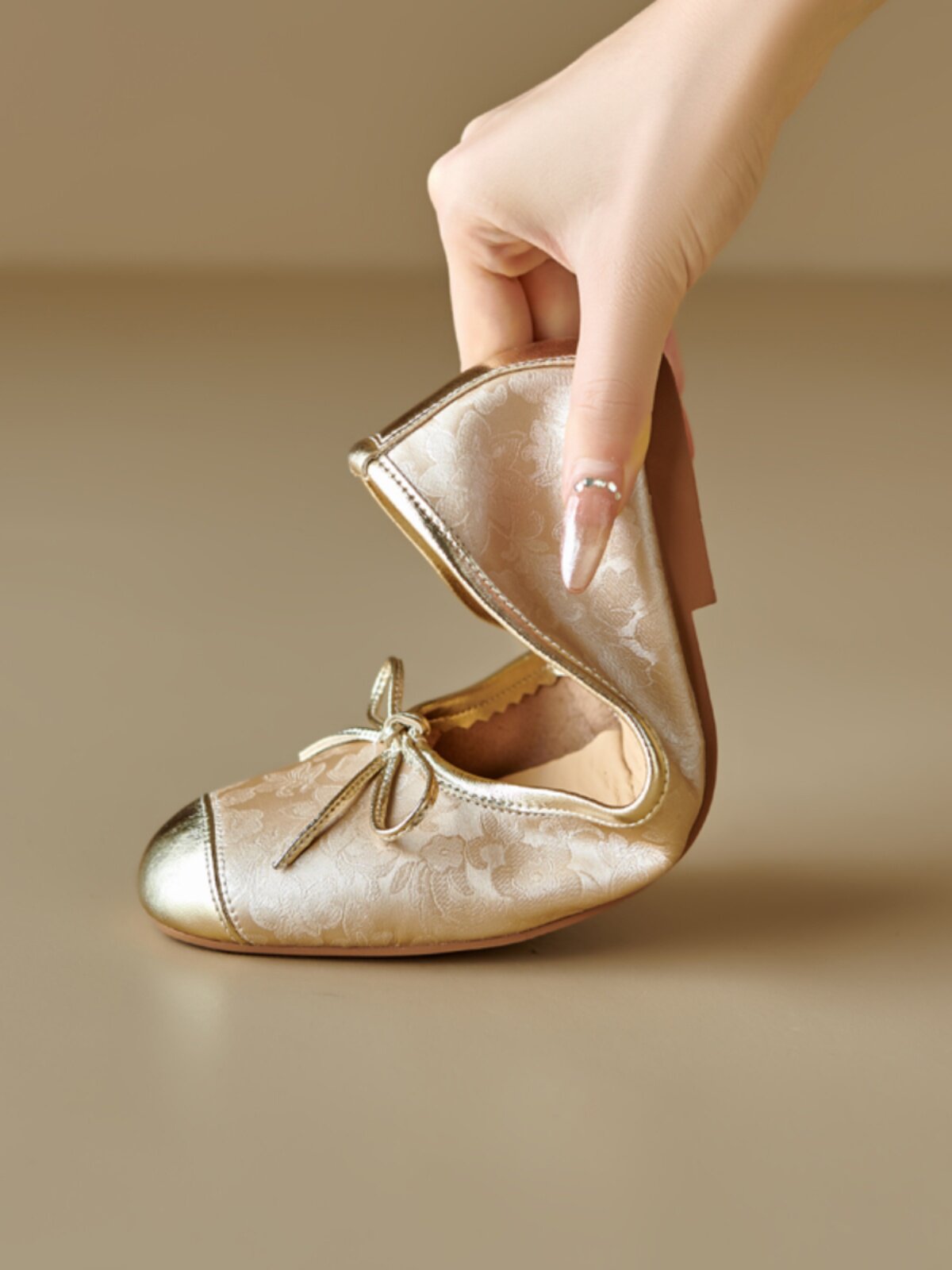 Gold floral ballet flats with metallic toe cap and bow, held by hand, close-up detail