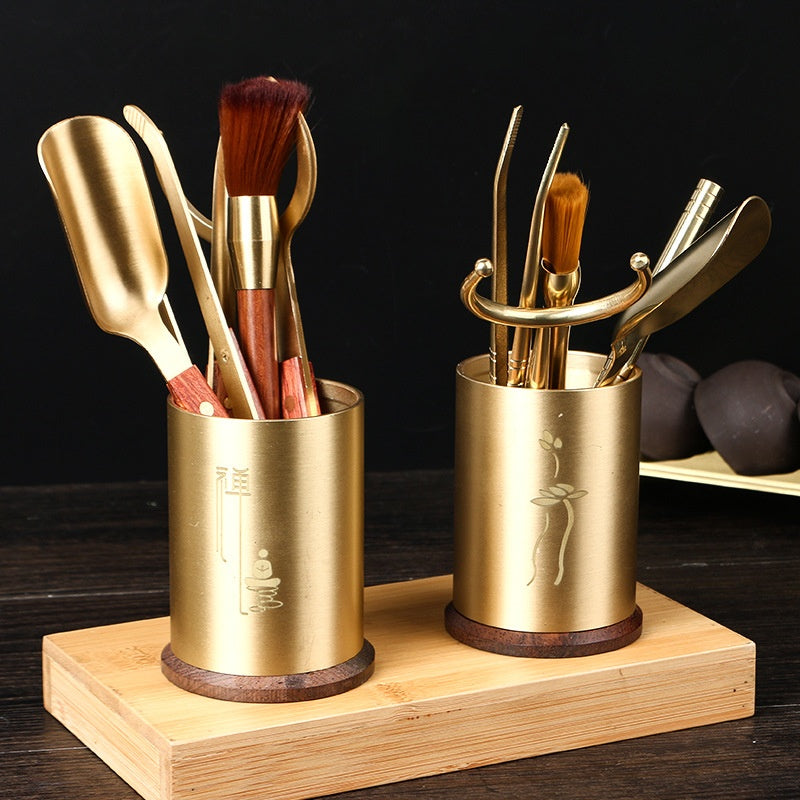 Tea ceremony tools with brass-coloured metal and dark wooden handles, arranged in two golden tubes on a wooden base, front