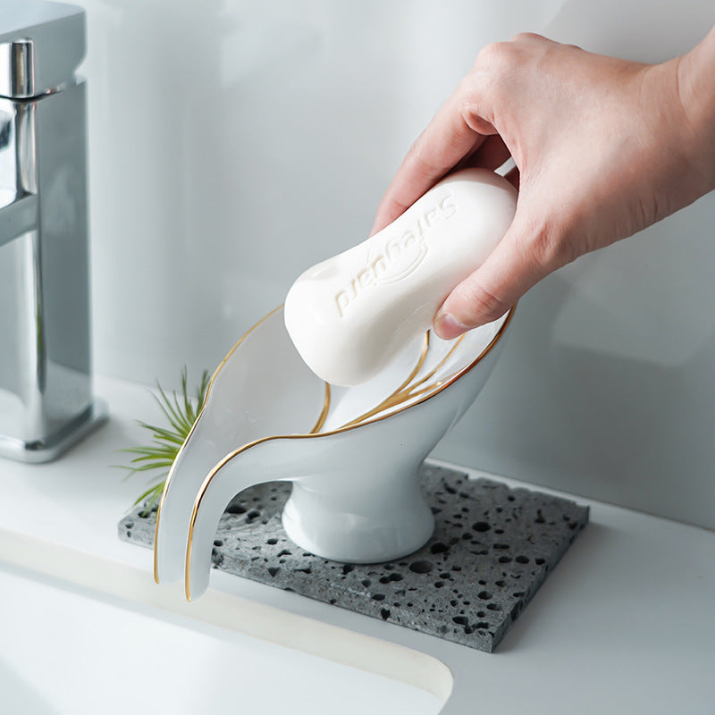 White ceramic leaf-shaped soap dish with gold-tone trim, being used to hold a bar of soap near a sink