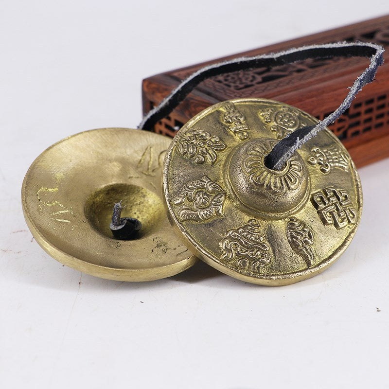 Tingsha cymbal pair with eight symbols, brass coloured, front and back view, joined by black cord, on white surface near
