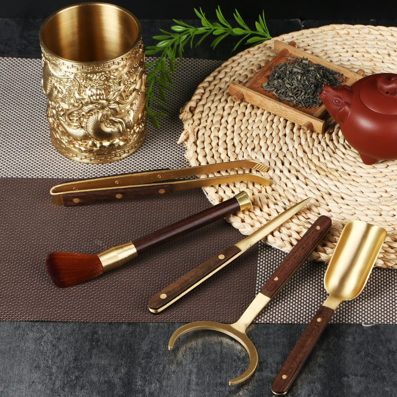 Tea ceremony tools with brass-coloured metal and red-brown wooden handles, laid flat on a textured mat near a teapot and tea