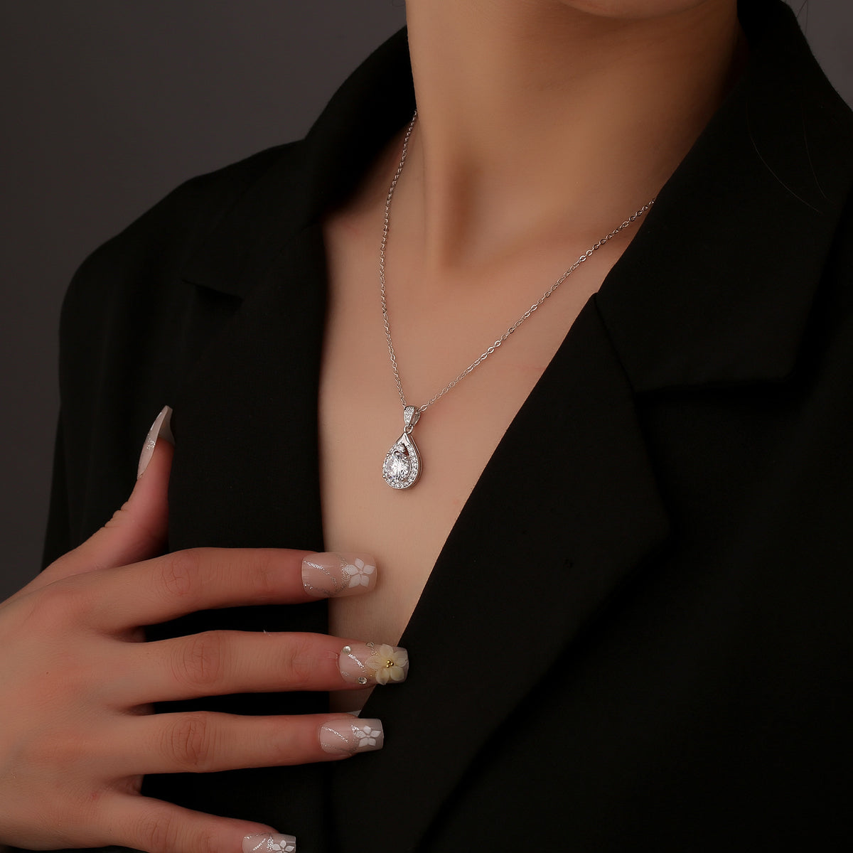 Druppelvormige hanger met ketting gedragen door een model in een zwarte blazer, hand rust bij het sleutelbeen
