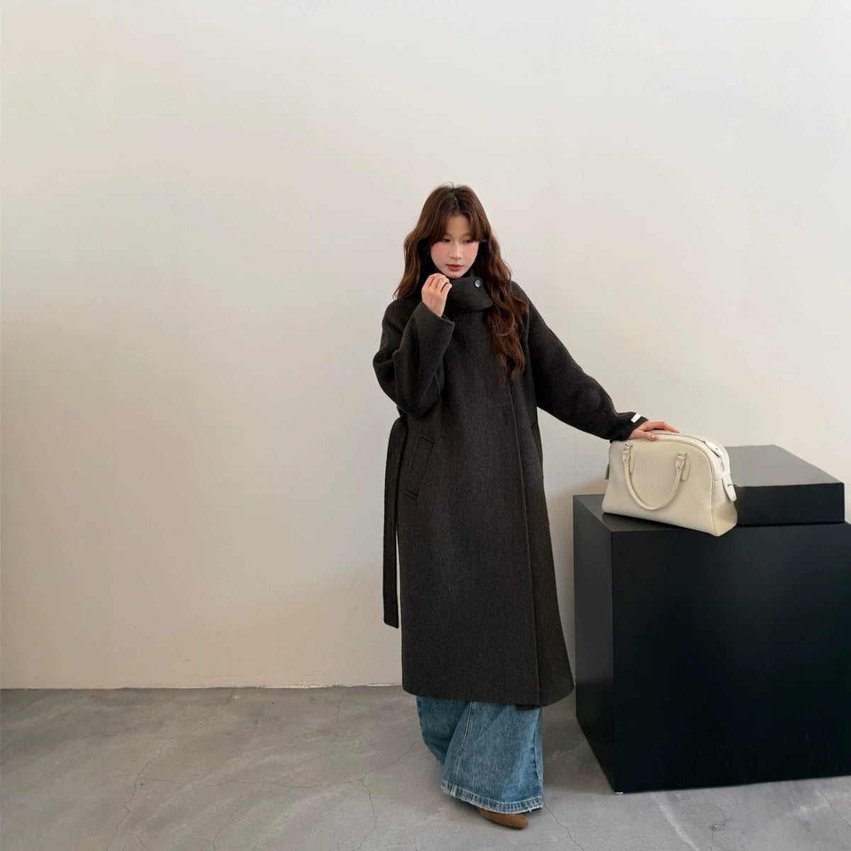 Charcoal grey long wool coat, side view on model resting a hand on a black plinth