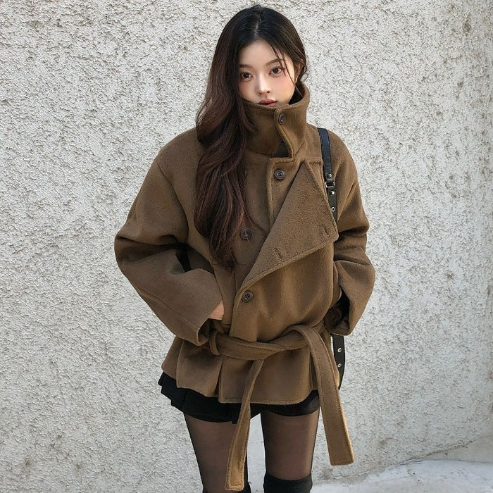 Brown cropped wrap jacket on model, front view, stand collar up, one hand in pocket, bag strap over shoulder