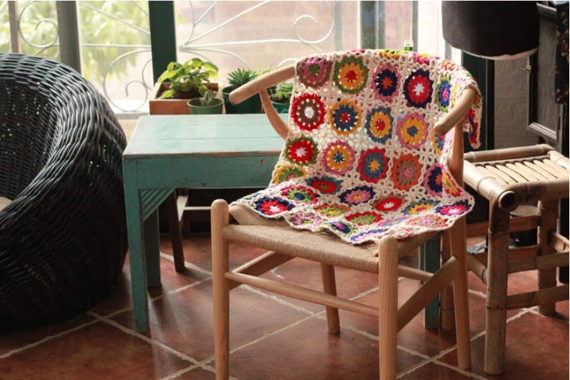 Crochet flower throw, multicolour, draped over wooden chair in bright room with plants