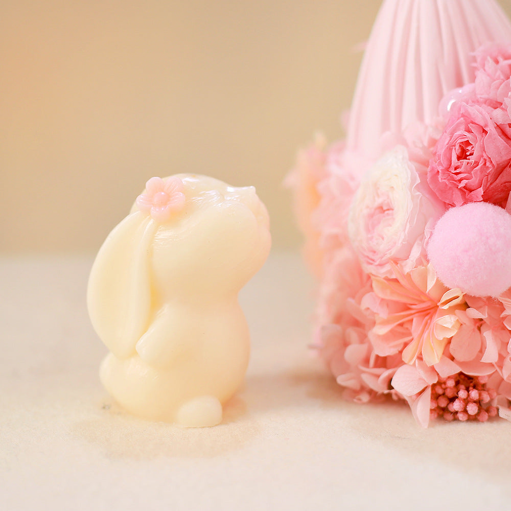 Close-up of a pale yellow rabbit-shaped candle with pink flower detail, next to pink floral arrangement on soft surface