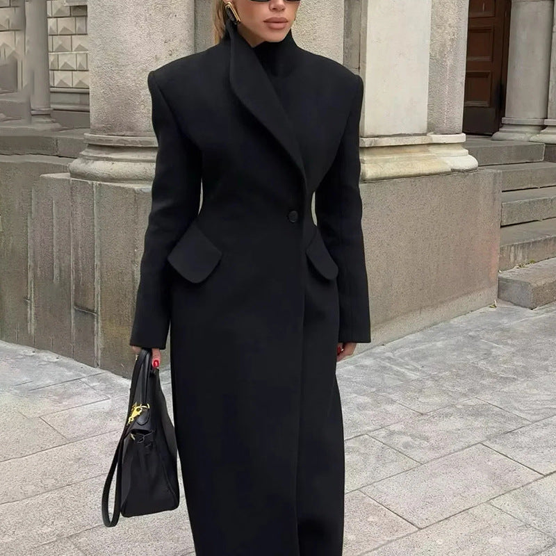 Model wearing a long black tailored coat with cinched waist and flap pockets, standing by stone columns