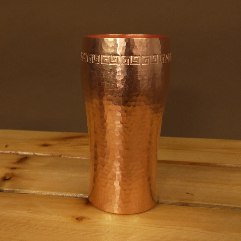 Curved hammered copper beer tumbler with Greek-key border, on wooden surface
