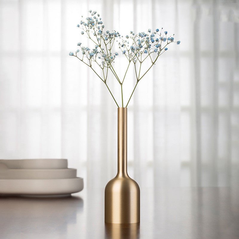 Brushed gold-tone metal bud vase with blue dried flowers on a reflective surface near stacked plates