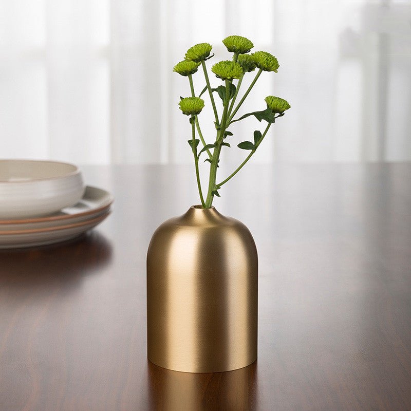 Brushed gold-tone metal bud vase with green flowers on a wooden table near plates