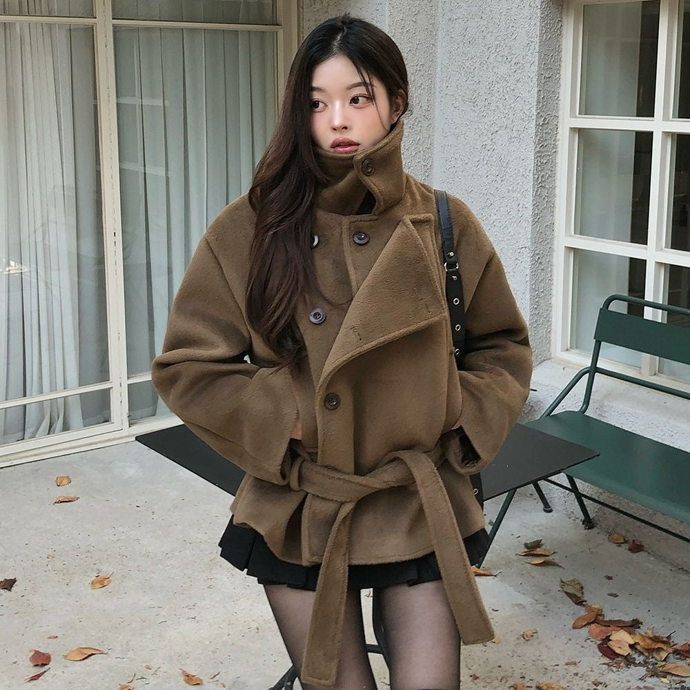 Brown cropped wrap jacket on model, front view, stand collar up, hands in side pockets, belt tied at waist