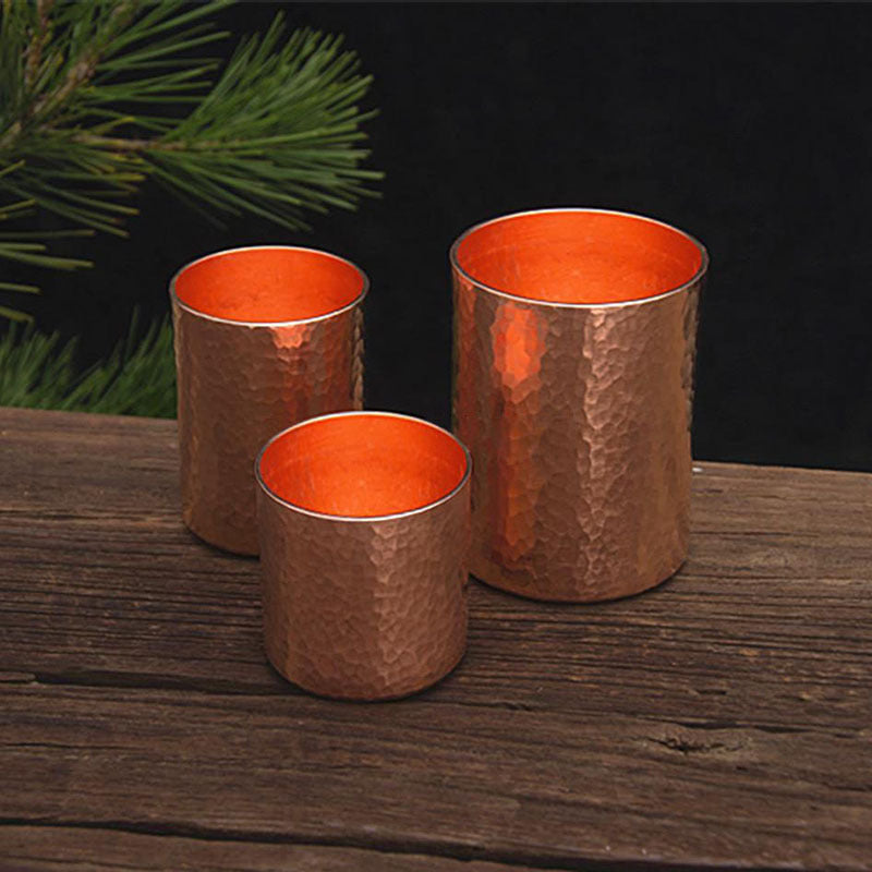 Three hammered metal tea cups, copper colour, arranged on wooden surface with pine branch in background